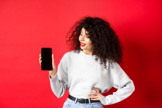 Portrait of stylish young woman with curly hair showing empty smartphone screen, demonstrating shopping app, standing on red background.