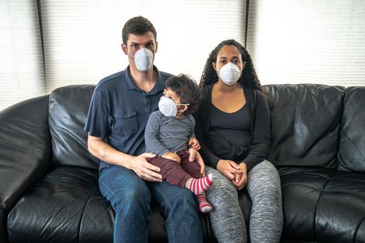 A young mixed family sits on a black leather sofa holding their son and wearing a dust face mask over their faces in hopes of preventing getting sick or ill from caronavirus or COVID-19.