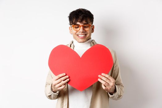 Handsome and stylish man in sunglasses, prepare surprise postcard for girlfriend on Valentines day, holding big red heart cutout and smiling happy, standing over white background.