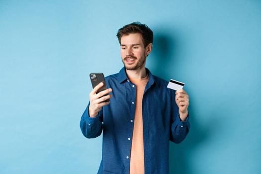 E-commerce concept. Image of handsome caucasian man paying online, looking at mobile phone screen and holding plastic credit card, blue background.