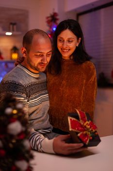 Husband surprising girlfriend with xmas present gift with ribbon on it celebrating christmas holiday in x-mas decorated kitchen. Happy joyful family enjoying winter season together