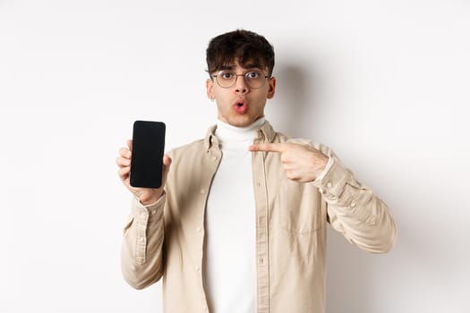 E-commerce concept. Portrait of young man pointing at mobile phone screen, showing advertisement online, standing on white background.