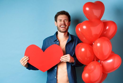 Heartbroken man crying of breakup of valentines day, holding red heart cutout and standing near romantic balloons over blue background.