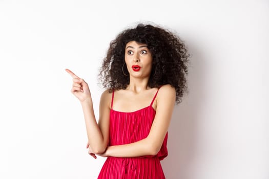 Excited girl with curly hair, wearing red dress, pointing finger left and looking amazed, showing cool promo, standing on white background.