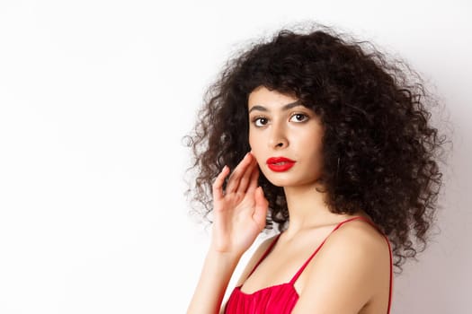 Close up of elegant woman with curly hair and red lips, looking at camera and touching face, standing in red dress on white background.