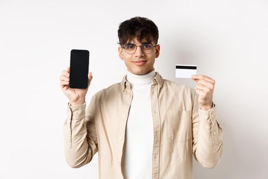 Online shopping. Young modern guy showing plastic credit card and empty smartphone screen, demonstrate account, standing on white background.