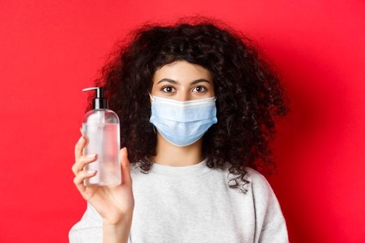 Covid-19, pandemic and quarantine concept. Young woman in medical mask showing bottle of hand sanitizer, demonstrate antiseptic, red background.