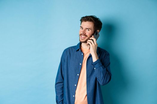 Modern smiling guy talking on mobile phone, looking happy at camera, standing on blue background.