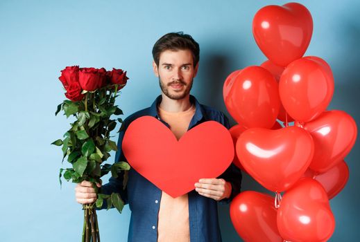 Valentines day romance. Young man with bouquet of red roses and heart balloons smiling, bring presents for lover on valentine date, standing over blue background.