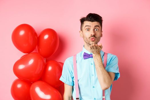 Valentines day concept. Attractive and funny man in bow-tie, blowing air kiss to lover, standing near pink background and red hearts balloons.