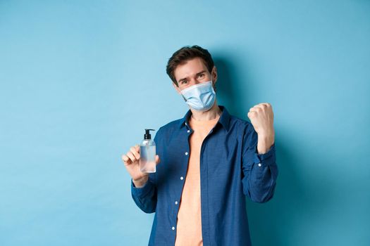 Coronavirus, quarantine and social distancing concept. Cheerful guy say yes and raise fist up while showing hand sanitizer, recommending product, blue background.