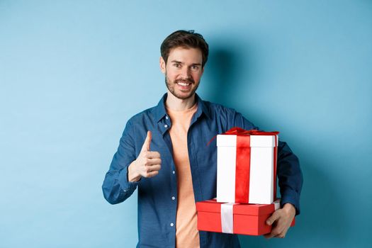 Smiling man holding romantic gifts and showing thumbs-up, celebrating Valentines day, buying presents for lover, standing over blue background.