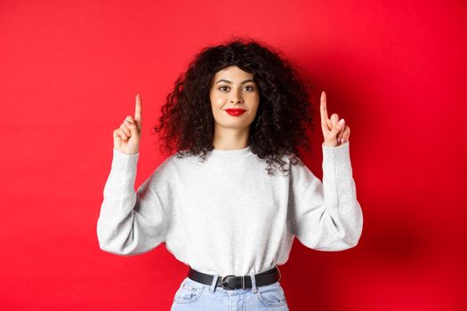 Image of cheerful smiling woman with curly hairstyle and red lips, pointing fingers up at empty space, showing advertisement, standing in casual clothes on studio background.