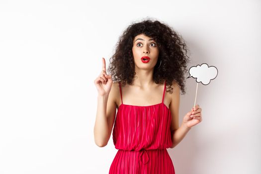 Excited fashionable woman in red dress pitching an idea, raising finger in eureka sign, holding thought cloud, standing on white background.