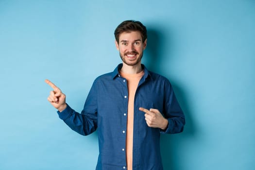 Happy smiling man pointing fingers left at logo, showing advertisement, standing on blue background.