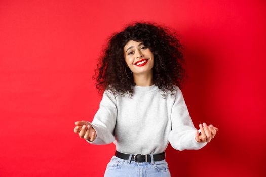 Pretty woman with curly haircut, extending hands and smiling with love and tenderness, inviting to come closer, reaching arms for hug, taking something into her arms, red background.