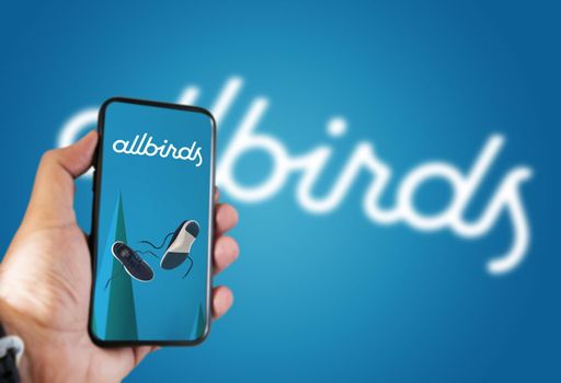 San Francisco, USA, October 2021: hand holding a phone with the Allbirds mobile app on the screen and the logo blurred on a blue background. Allbirds designs and sells footwear and apparel