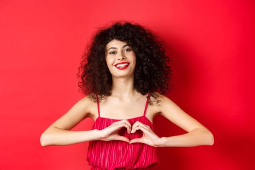 Lovers day. Beautiful woman celebrating valentines, showing heart sign and smiling, standing in romantic red dress on studio background.