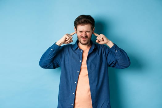 Annoyed young man block sound, shut ears with fingers and grimacing, disturbed by loud music, standing on blue background.