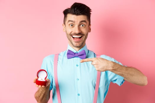 Valentines day. Cheerful young man going to make proposal, pointing finger at engagement ring and smiling excited, standing on romantic pink background.