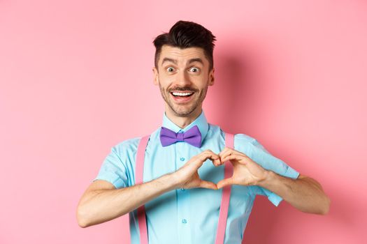 Passionate guy in funny bow tie saying I love you, showing heart gesture on Valentines day and smiling, expressing sympathy to lover, standing over pink background.