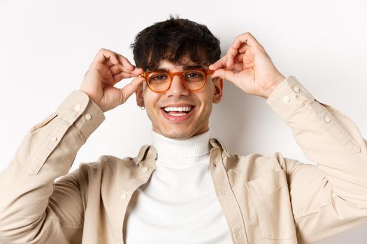 Close-up of stylish hipster guy trying eyewear at optician store, put on glasses and smiling, standing on white background.