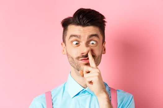 Image of funny caucasian guy picking nose and squint eyes, making dumb face, standing on pink background.