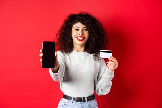 Happy good-looking girl showing plastic credit card and empty smartphone screen, demonstrate account or application, standing on red background.