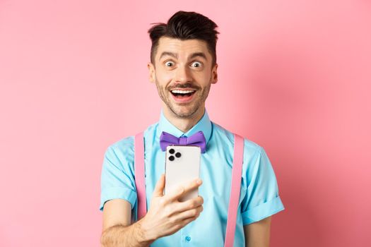 Image of excited man checking out promo offer online, holding smartphone and stare surprised at camera, standing happy on pink background.