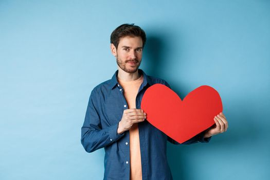 Handsome bearded guy showing valentines day cutout and smiling, standing in casual clothes over blue background. Copy space