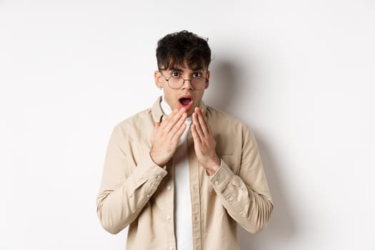 Portrait of handsome guy student looking shocked, hear gossip and stare with disbelief, covering opened mouth with hands, standing on white background.