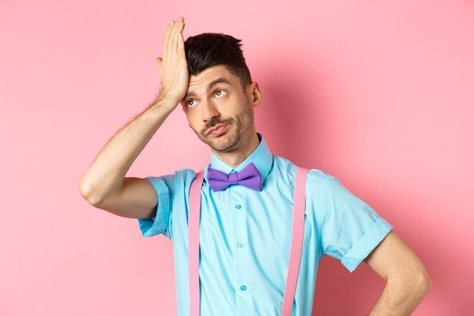 Annoyed guy with suspenders and bow-tie, facepalm with eyes rolled up, standing irritated by something stupid, standing over pink background.