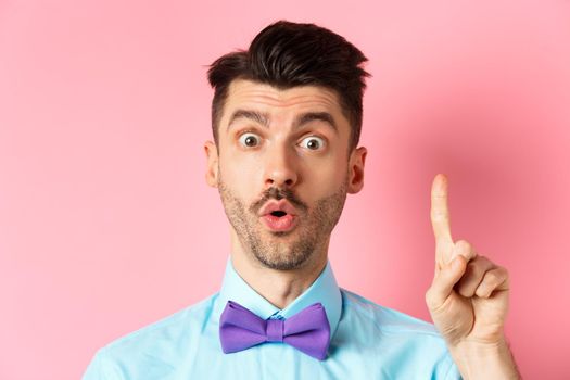 Close-up of excited funny guy with moustache and bow-tie, pitching an idea, raising finger to say suggestion, have plan, standing over pink background.