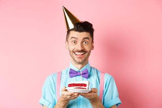Holidays and celebration concept. Cheerful young man celebrating birthday in party hat, holding b-day cake with candle and making wish, smiling happy at camera, pink background.