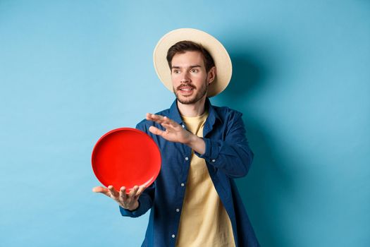 Happy smiling guy throwing frisbee, looking aside at friend, standing on blue background in straw hat.
