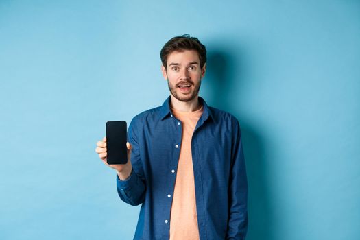 Happy caucasian guy showing empty mobile screen and smiling, standing on blue background.