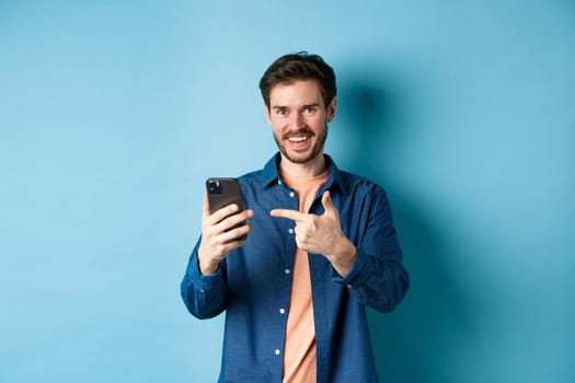 Handsome smiling man pointing finger at smartphone and looking pleased at camera, recommending app, standing on blue background.