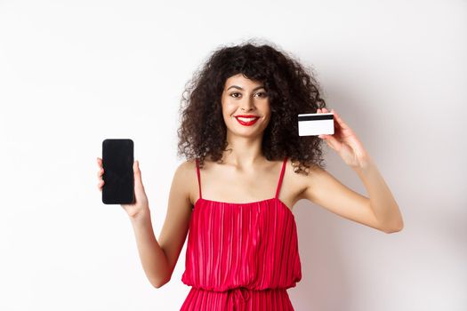 Online shopping concept. Elegant female model with curly hair, wearing red dress, showing plastic credit card with empty mobile phone screen, standing on white background.