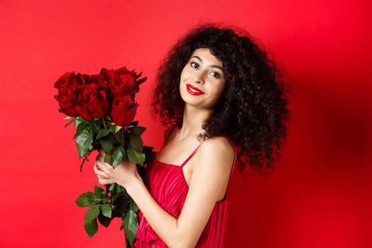 Happy beautiful woman in dress, holding flowers and smiling romantic, standing against red background.