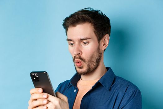 Image of impessed young man looking at smartphone and saying wow, checking out online offer, standing on blue background.