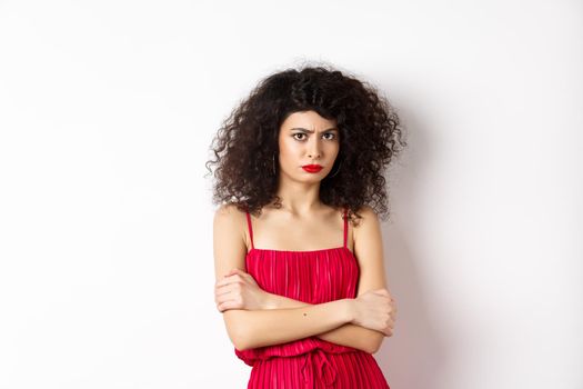 Sad and offended caucasian woman in red dress frowning, cross arms on chest and sulking, feeling mad at you, standing over white background.