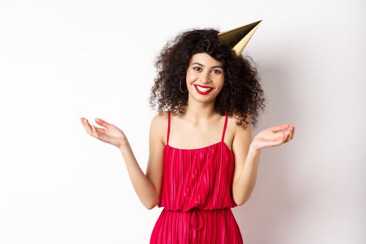 Beautiful caucasian woman in red dress, celebrating holiday, wearing party hat and smiling, standing on white background. Copy space