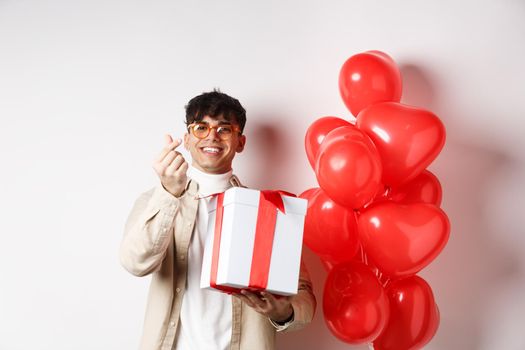 Valentines day and romance concept. Handsome young man showing surprise gift for lover and finger heart, smiling, standing near festive red balloons, white background.