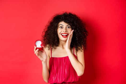 Image of surprised young woman with curly hairstyle, wearing red dress and lipstick, showing engagement ring, going to get married, standing on studio background.