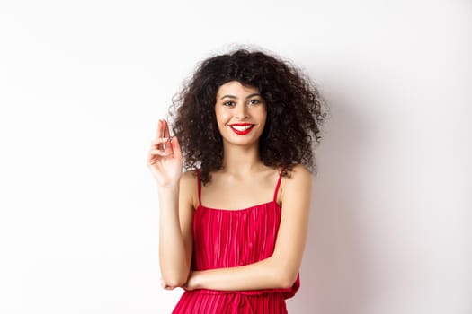 Beauty and fashion. Smiling woman with curly hair and makeup, wearing red dress, waving hand in greeting gesture, saying hello, standing on white background.