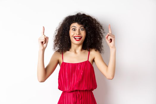 Happy elegant woman in red dress and makeup, smiling amused and pointing fingers up at logo, showing advertisement, standing over white background.