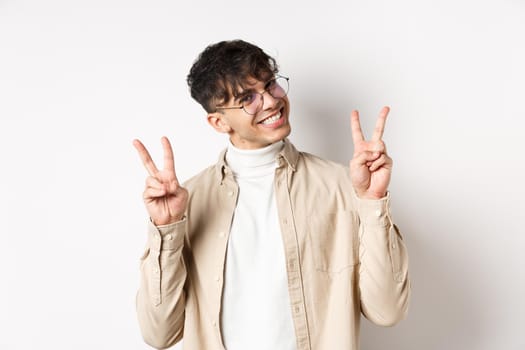 Real people. Handsome hipster guy in glasses showing peace gestures and smiling cute, standing on white background.