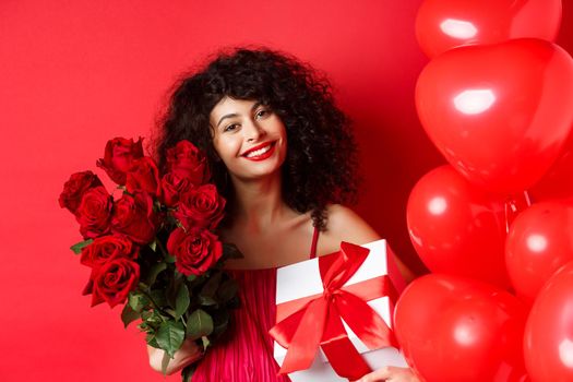 Romantic woman receive bouquet of flowers and gift on holiday, standing near heart balloons and red background, smiling grateful.