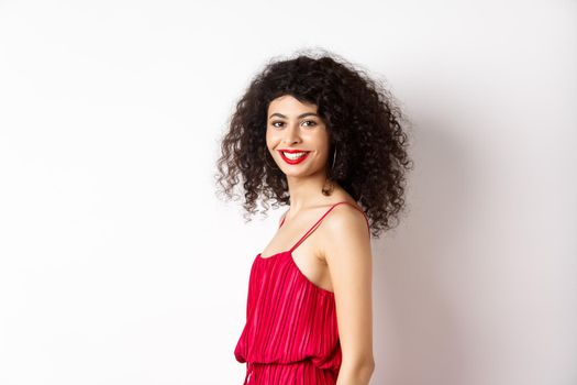 Profile of fashionable curly-haired woman in red dress, turn head at camera and smiling confident, standing over white background.