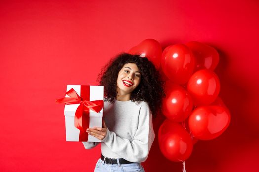 Holidays and celebration. Happy birthday girl holding gift and posing near party helium balloons, smiling excited at camera, red background.